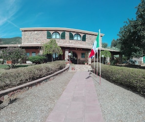 A stone building with a curved roof and arched windows, a paved pathway leading to the entrance, and a flagpole with a flag. - Hotel Janas (Tertenia)