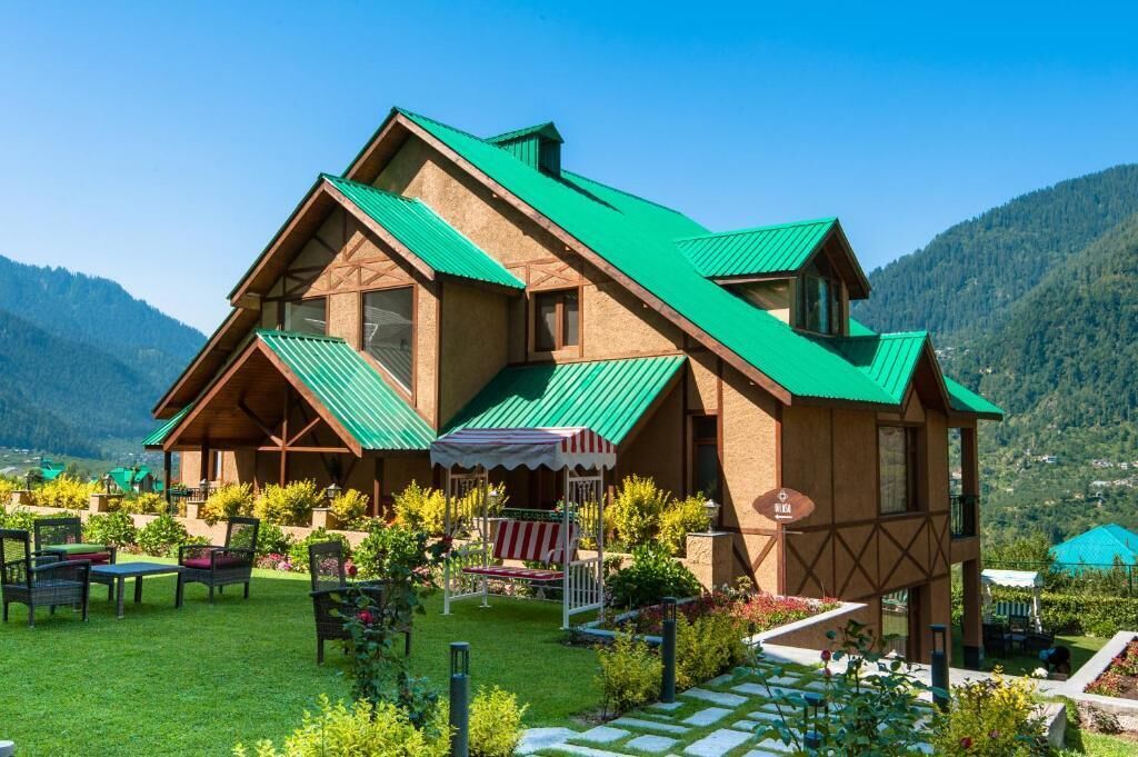 Um edifício em estilo chalé com telhado inclinado, rodeado por um jardim bem cuidado e uma paisagem montanhosa ao fundo.