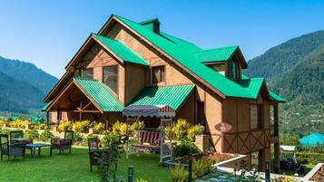 Um edifício em estilo chalé com telhado inclinado, rodeado por um jardim bem cuidado e uma paisagem montanhosa ao fundo.