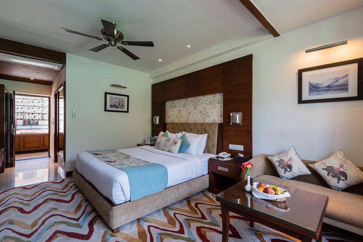 A hotel room with a large bed, a sofa, a coffee table, a ceiling fan, and a view of the outside.