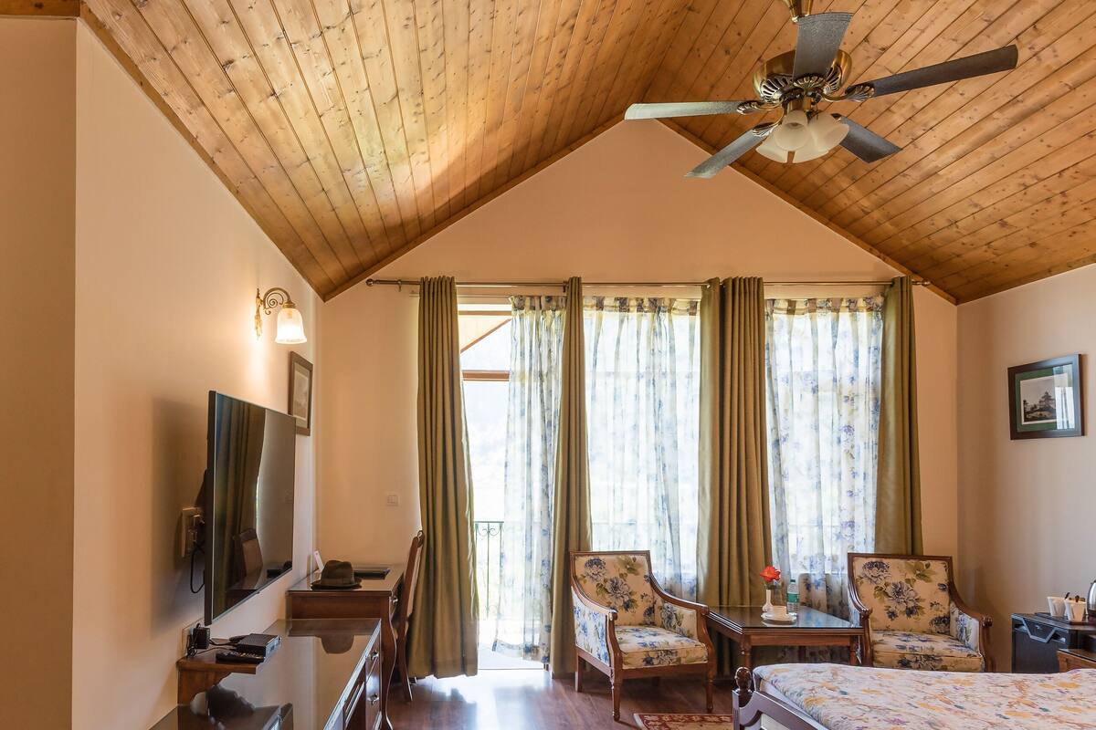 A bedroom with a wooden ceiling, a bed, two chairs, a desk, and a television.