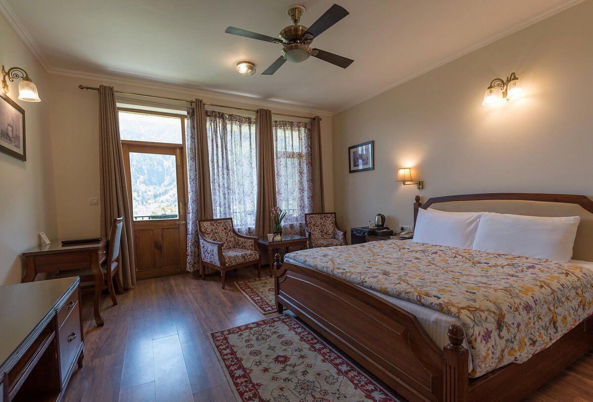 A bedroom with a wooden bed, a desk, two chairs, a nightstand, and a ceiling fan.