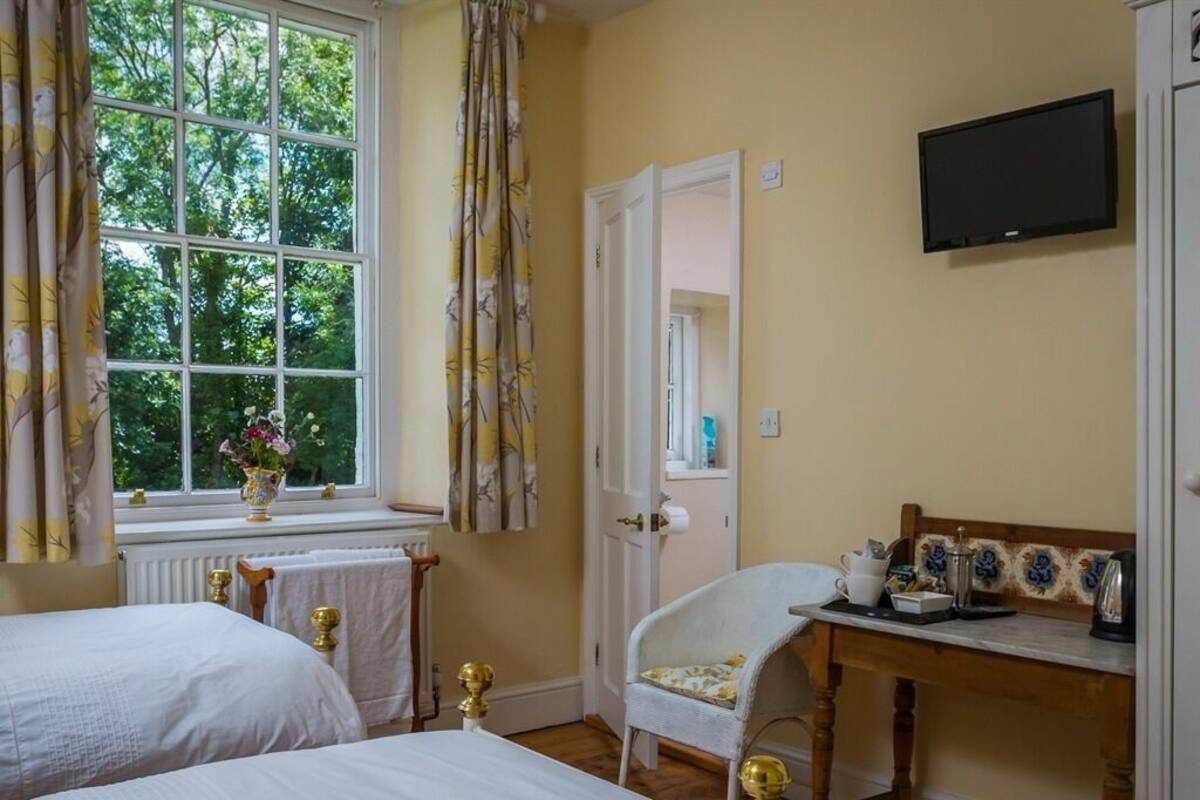 A bedroom with two beds, a window with curtains, a chair, a table with a kettle, and a television on the wall.