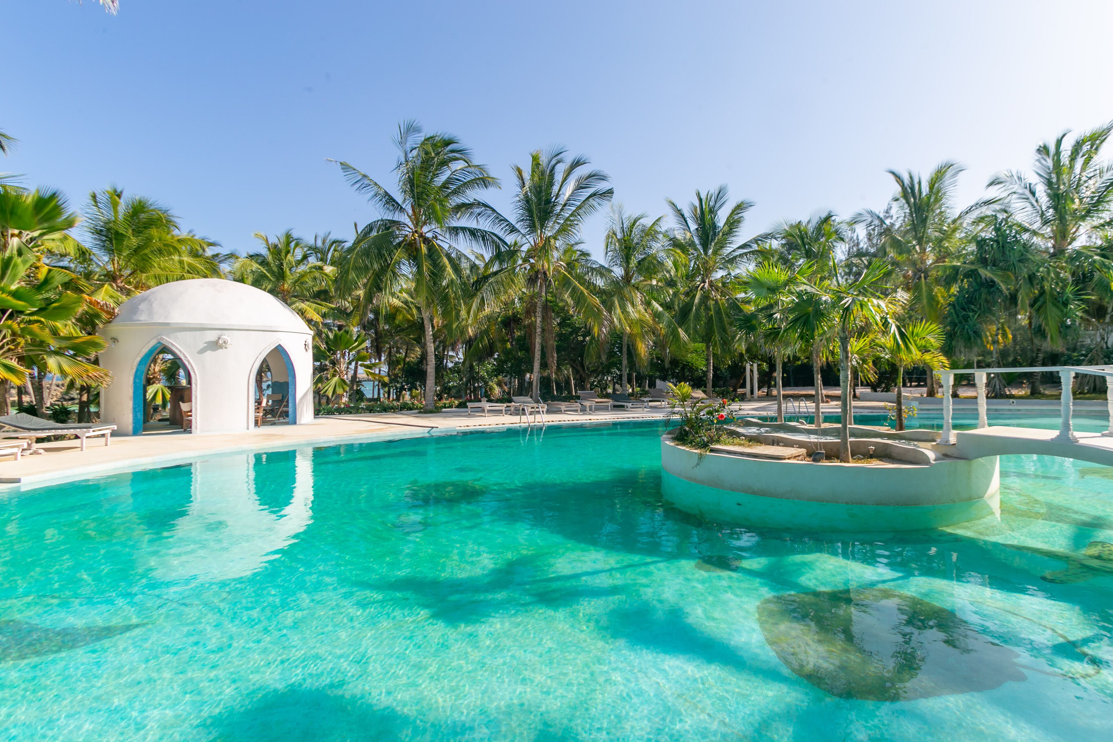 A clear blue pool with a small pavilion and palm trees in the background.