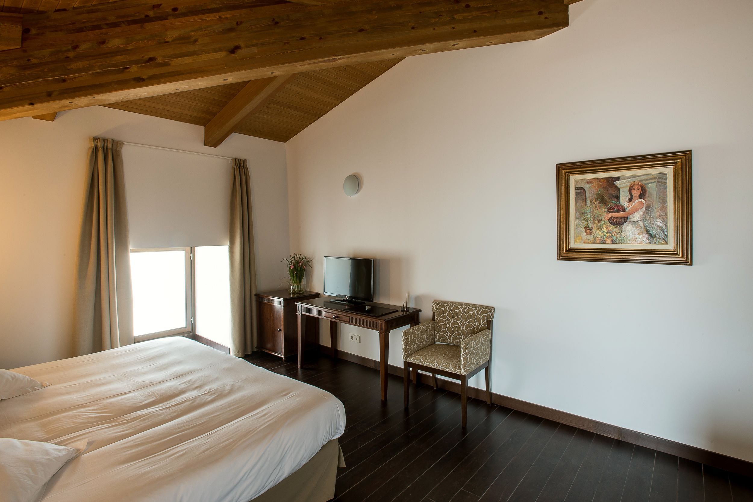 A hotel room with a bed, a desk with a television, a chair, and a painting on the wall.