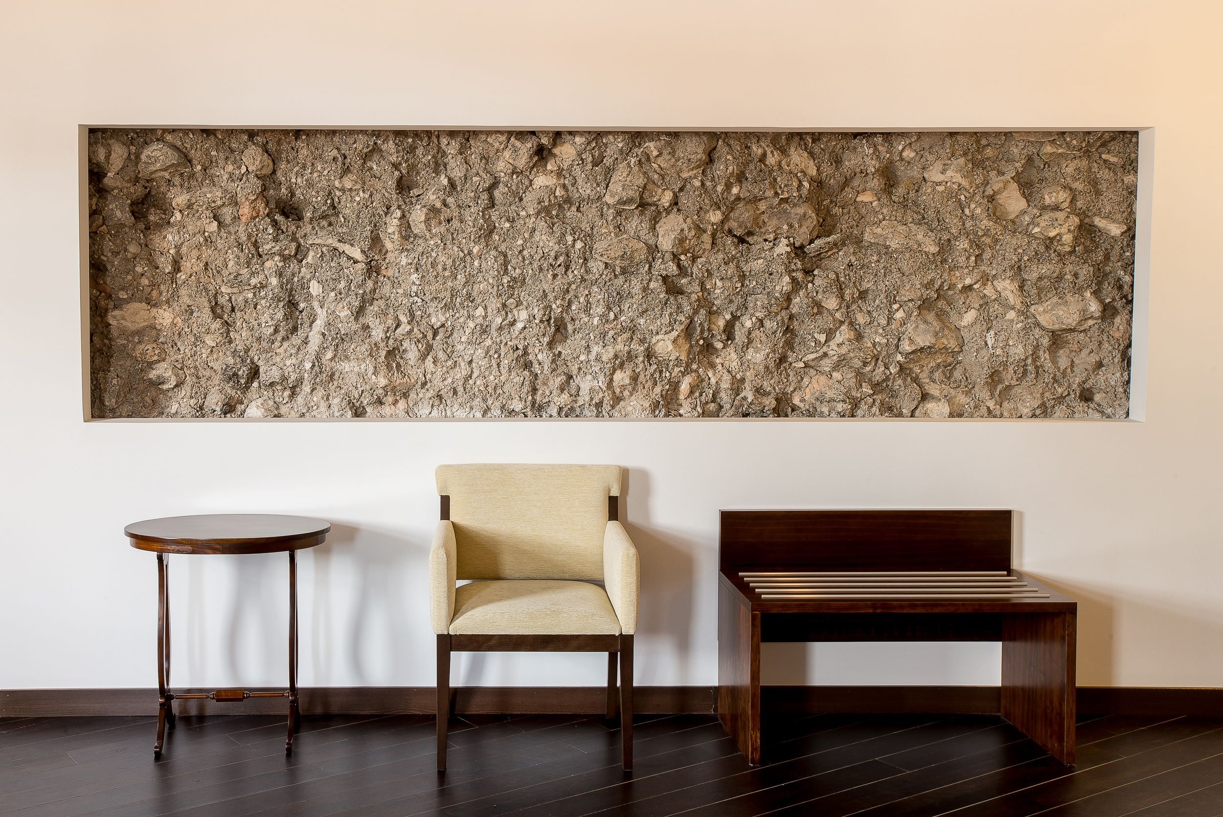 Una habitación con un cuadro de pared grande y con textura, una mesa redonda de madera, un sillón color beige y un banco de madera.