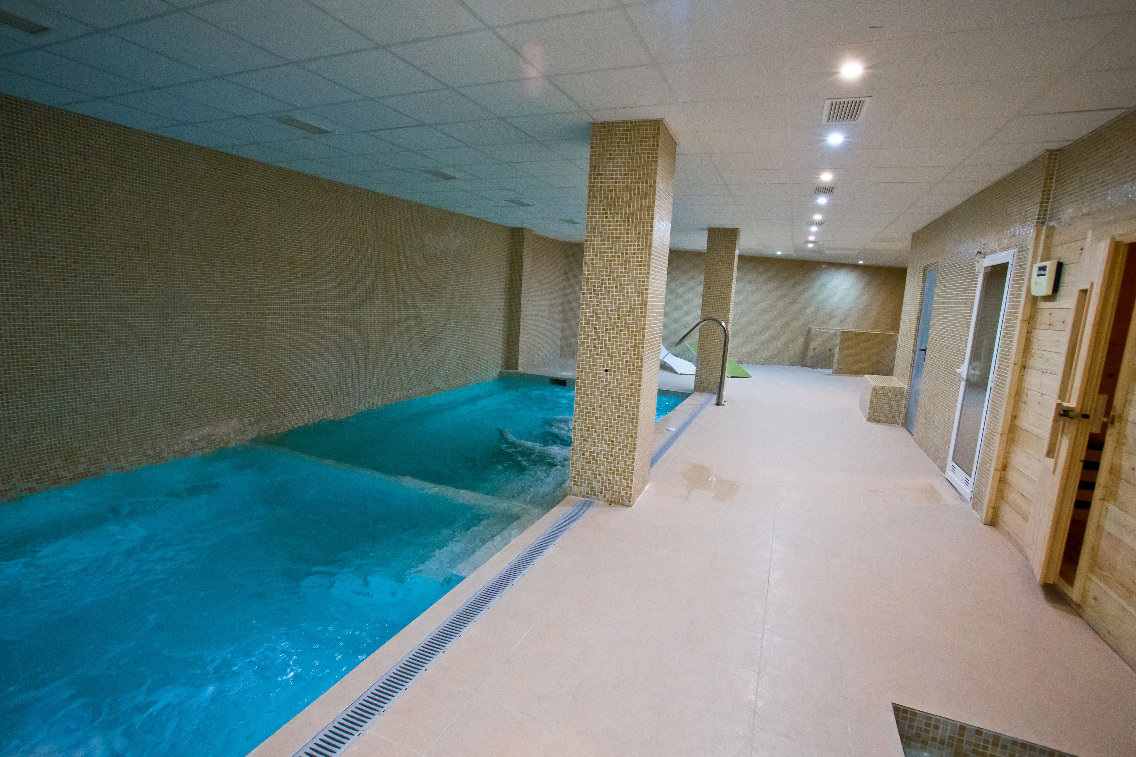 An indoor swimming pool with tiled walls and a tiled floor, a central column, and a door on the right.