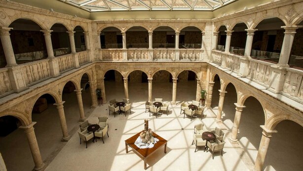 Un amplio patio con varios niveles de arcos y columnas, amueblado con mesas y sillas, y una pieza decorativa en el centro.