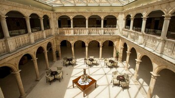 Un amplio patio con varios niveles de arcos y columnas, amueblado con mesas y sillas, y una pieza decorativa en el centro.