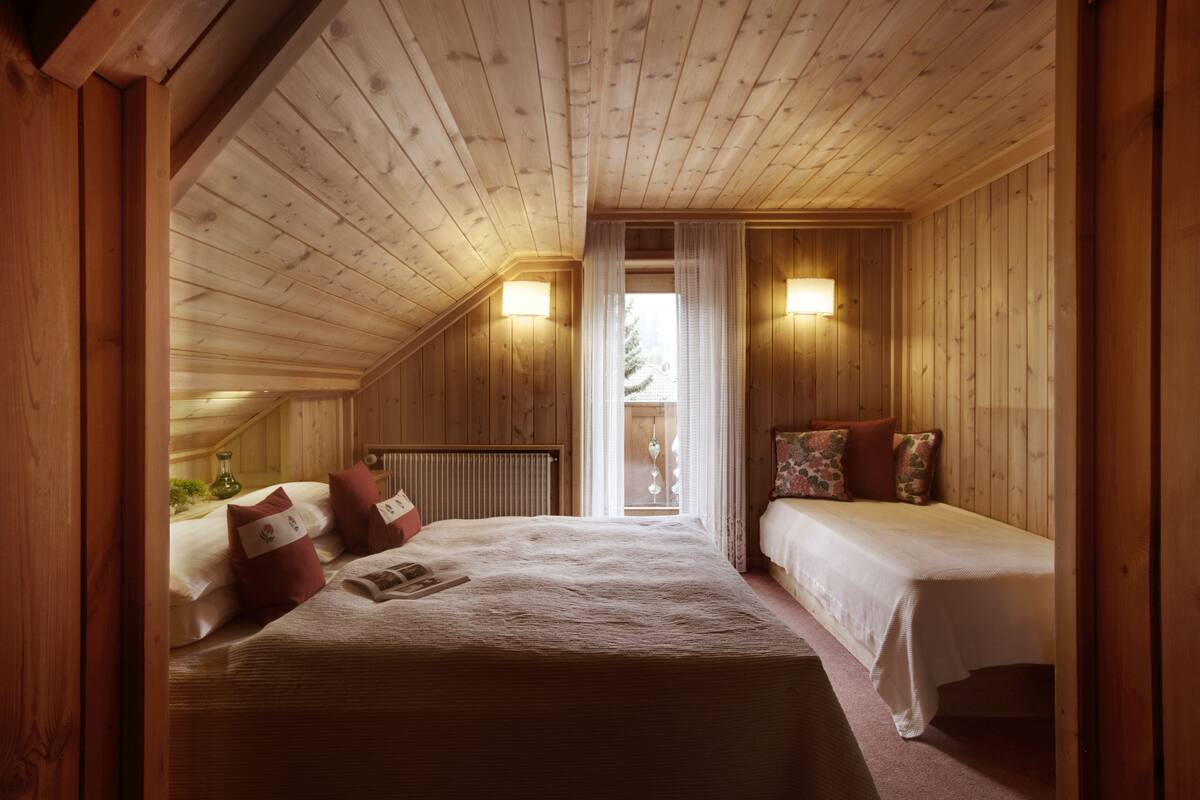 A cozy attic bedroom with wooden walls and sloped ceilings, featuring two beds, a radiator, and a window with curtains.