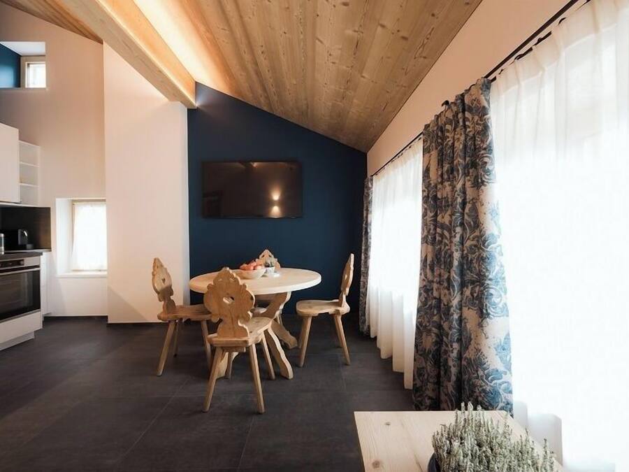 A modern kitchen with built-in appliances, a dining area with wooden chairs, and a large window with patterned curtains.