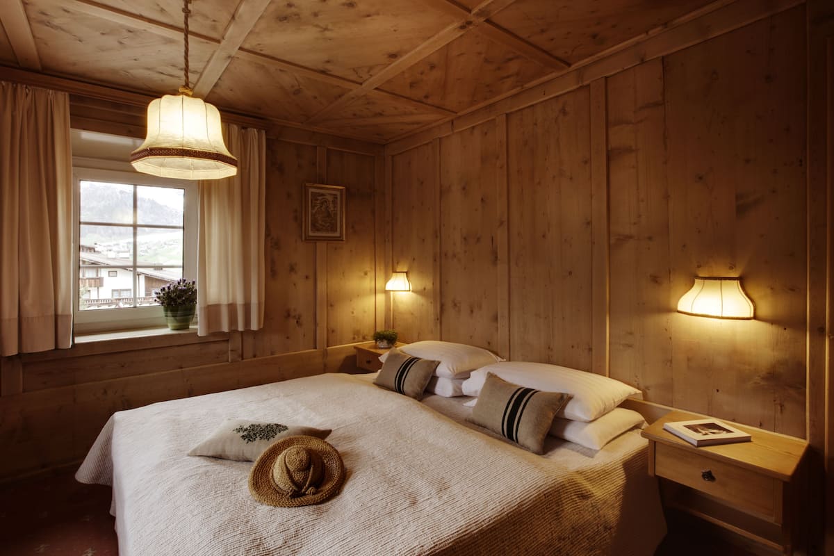 A wooden room with a bed, two bedside lamps, a window with curtains, and a framed picture on the wall.