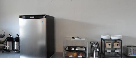 A kitchenette with a stainless steel refrigerator, a coffee machine, a toaster, and a small glass bowl on a white countertop.
