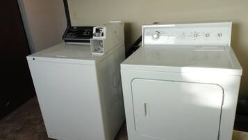 Deux machines à laver à chargement frontal, de couleur blanche, se trouvent dans une pièce dont le mur est uni et où se trouve une porte sur le côté gauche.