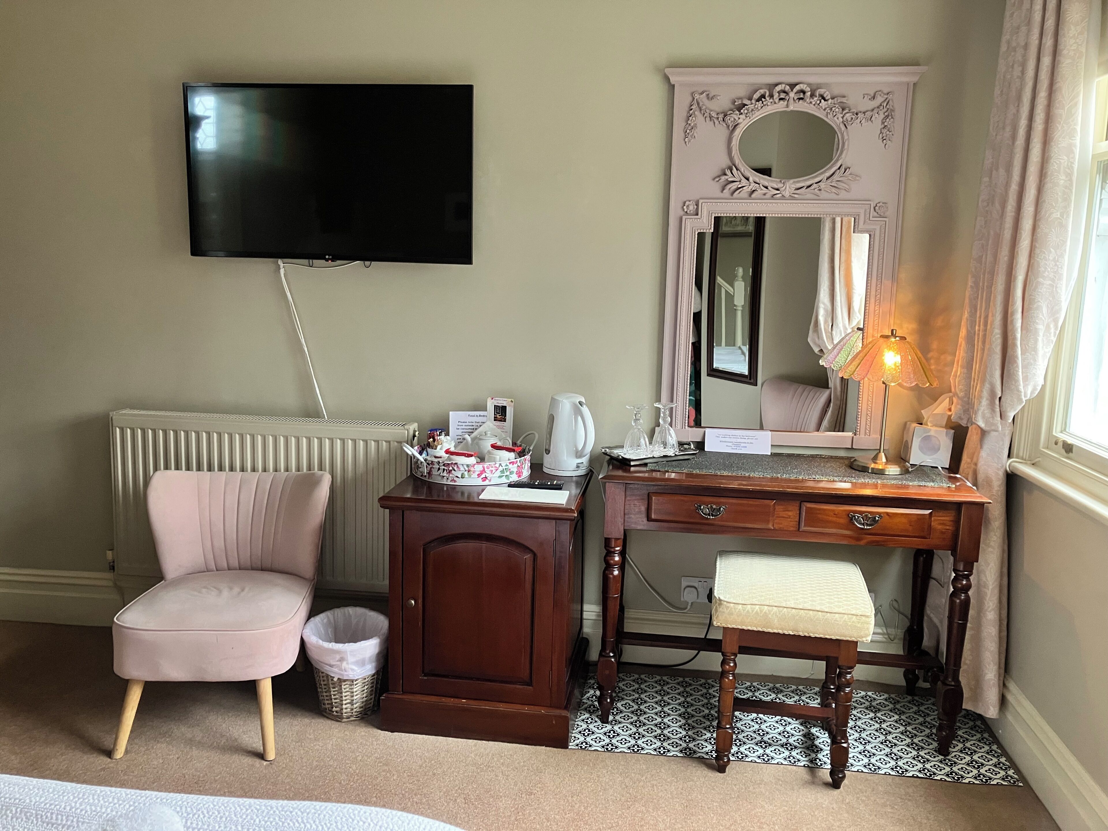 A hotel room with a television, a vanity with a mirror, a chair, a desk, and a bedside table.