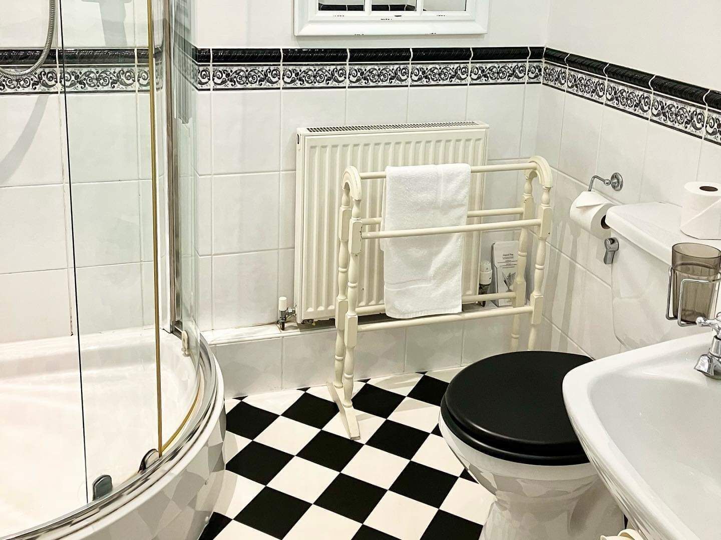 Een badkamer met een zwart-wit schakeerd patroon, een witte radiator, een glazen douchecabine en een witte wastafel met een glazen houder.