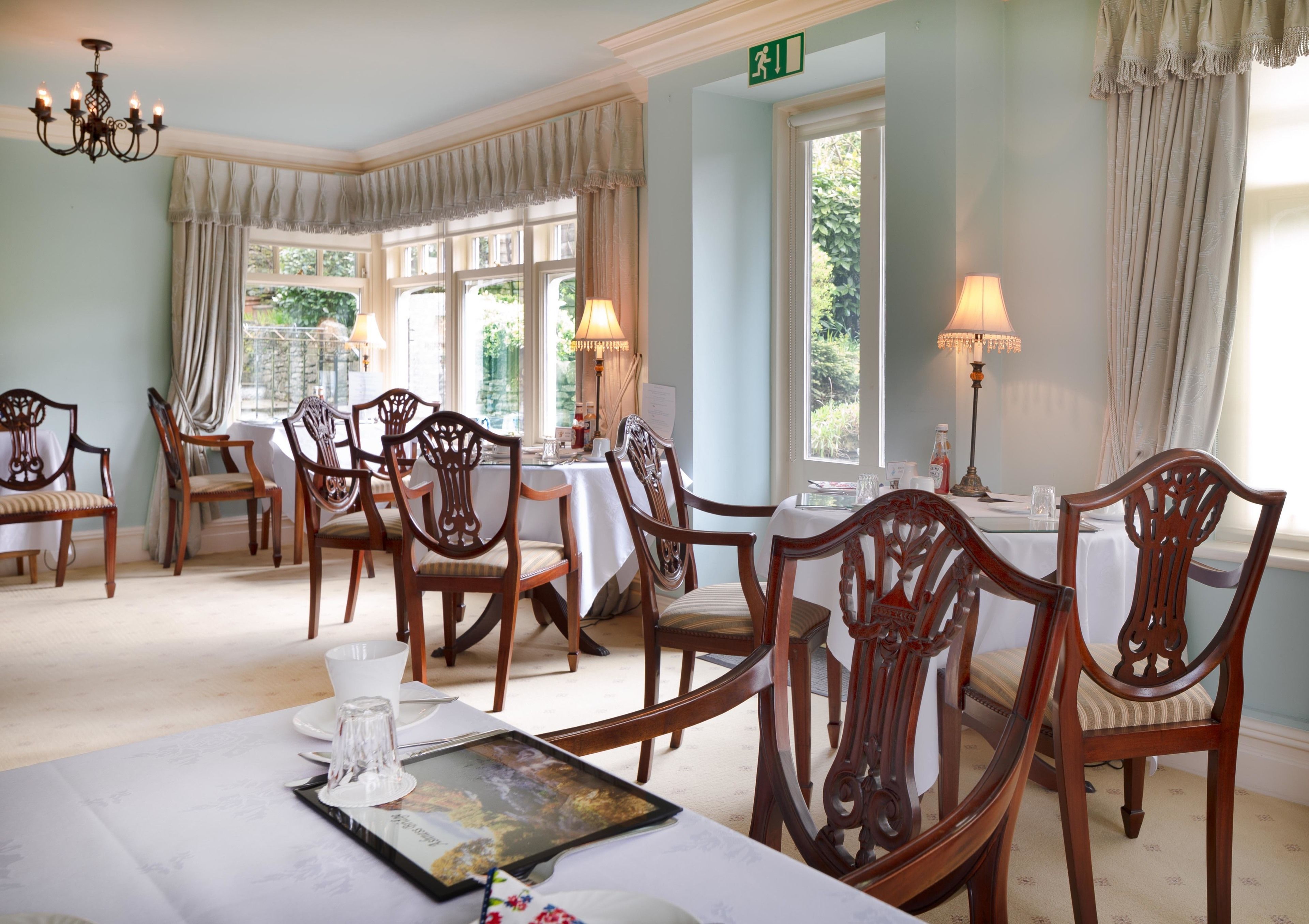 A dining area with wooden chairs and tables, large windows with curtains, and a chandelier.