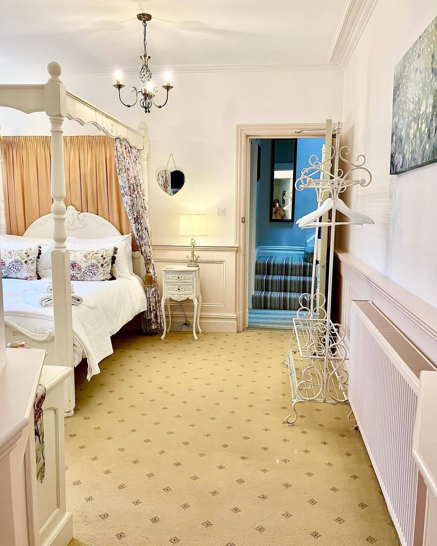 A bedroom with a four-poster bed, a bedside table, a lamp, and a hanging mirror.