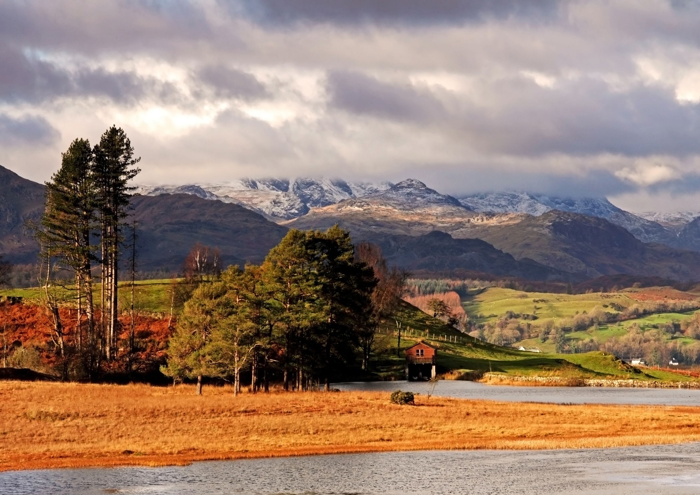 A rural landscape with a house near a body of water, surrounded by mountains and autumn foliage.