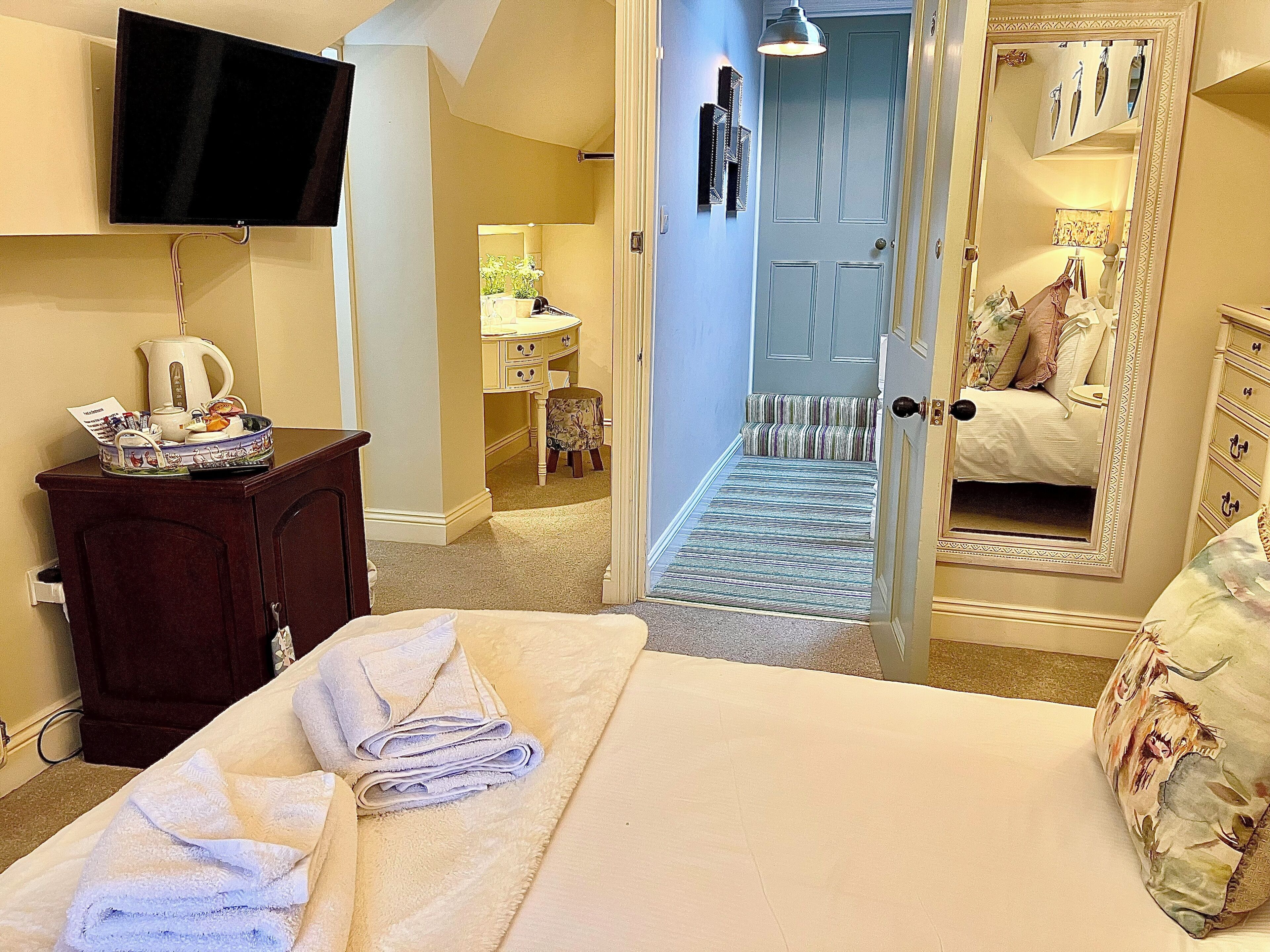 A hotel room with a bed, a TV mounted on the wall, a desk with a kettle, and a bathroom visible through an open door.