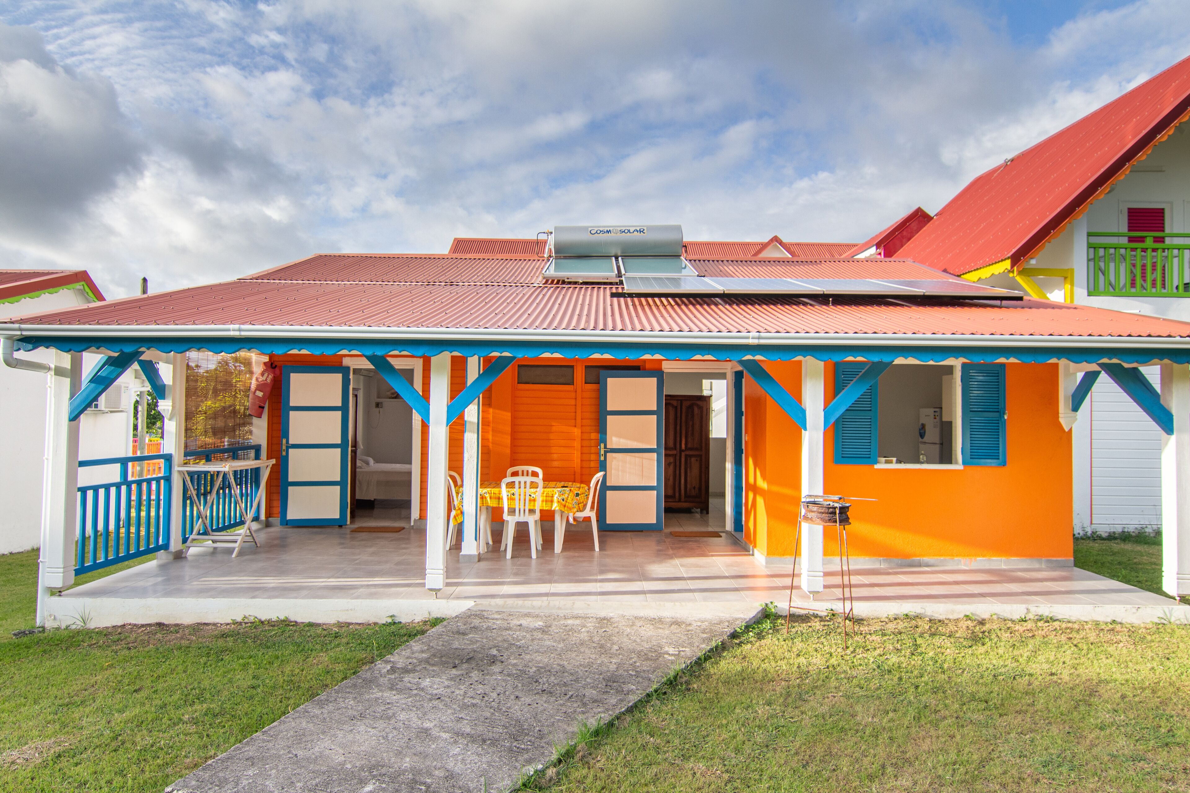 Une maison aux couleurs vives, avec un toit rouge, une façade bleue et blanche, et une véranda abritée avec des chaises.