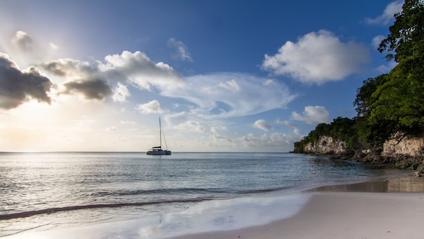 A beach with a sailboat in the water, a sandy shore, and a rocky cliff with vegetation. - Au Jardin Debeauséjour (Capesterre-de-Marie-Galante)