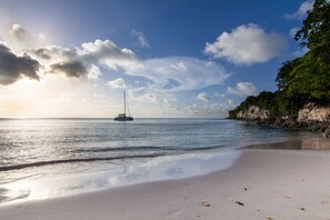 A beach with a sailboat in the water, a sandy shore, and a rocky cliff with vegetation. - Au Jardin Debeauséjour (Capesterre-de-Marie-Galante)