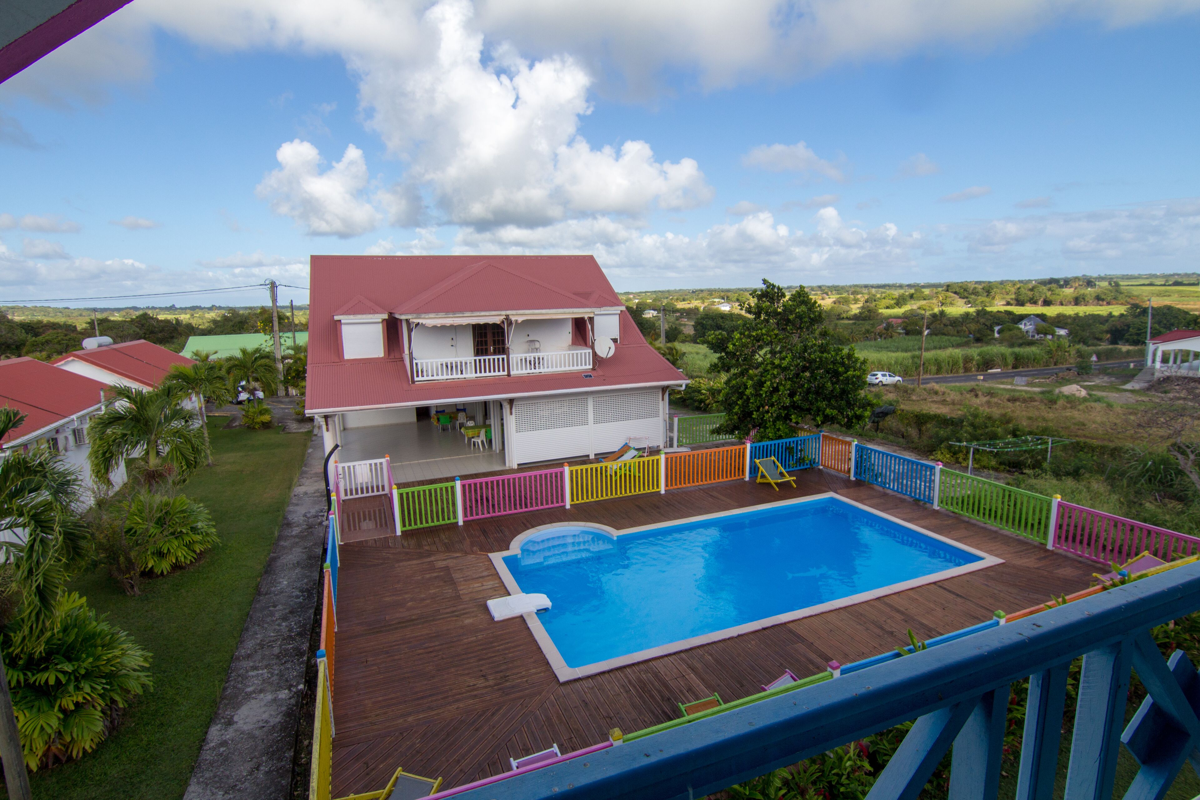 A house with a swimming pool surrounded by colorful fences, a balcony, and a view of the countryside.