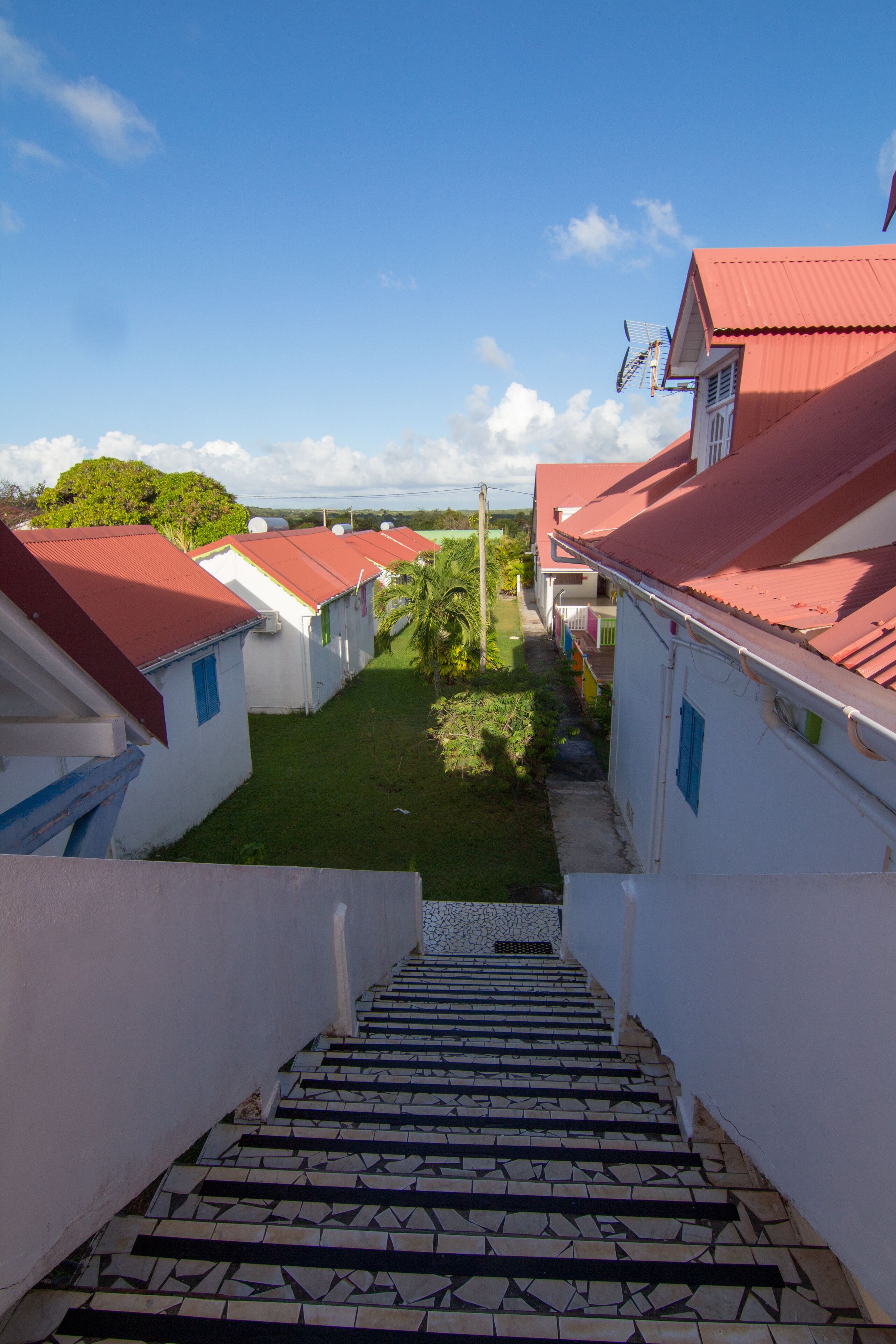 A staircase leading to a row of white buildings with red roofs, a clear sky, and a grassy area with trees.