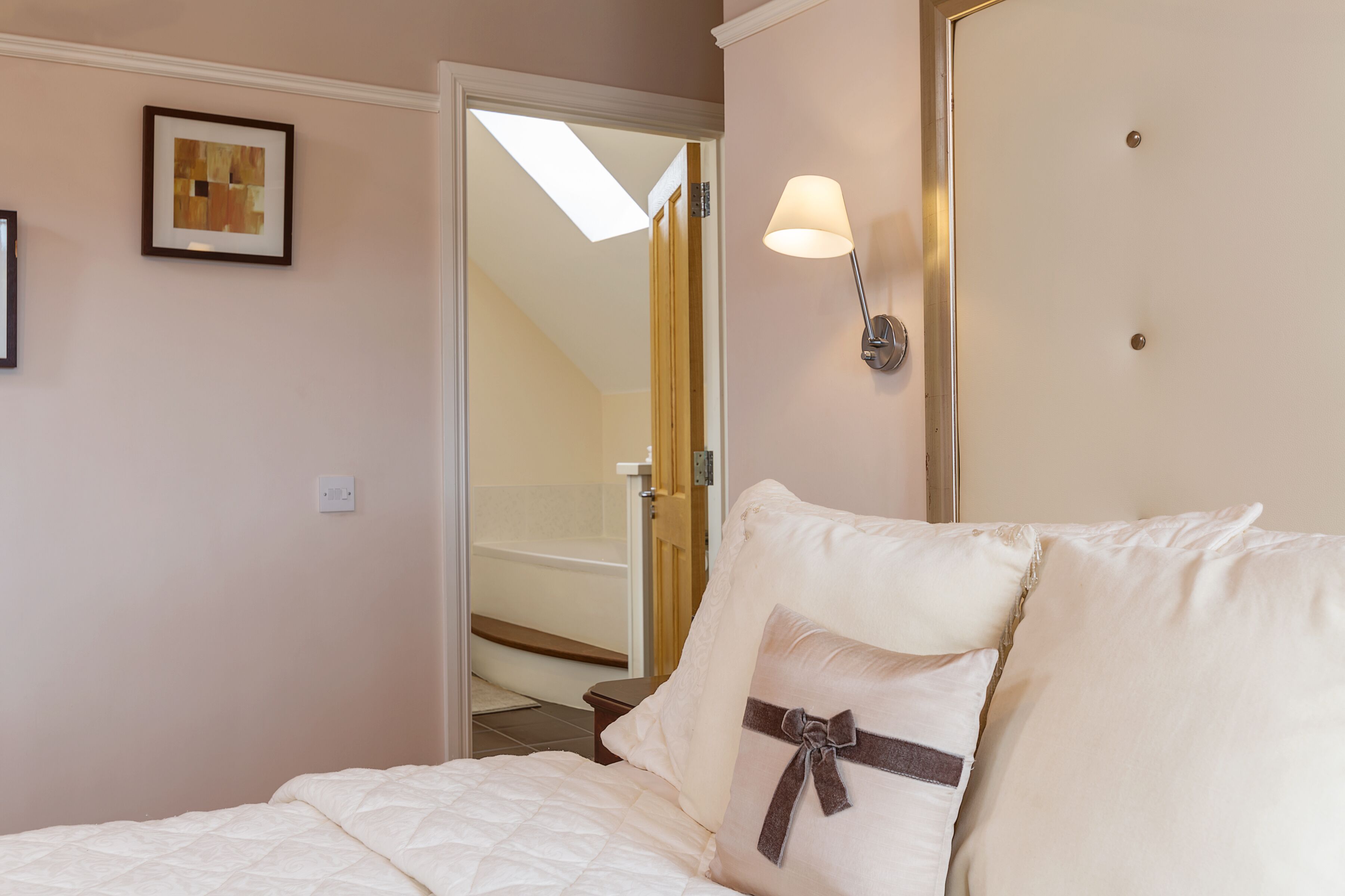 A bedroom with a bed, a nightstand, a wall-mounted lamp, a framed picture, and a door leading to a bathroom.