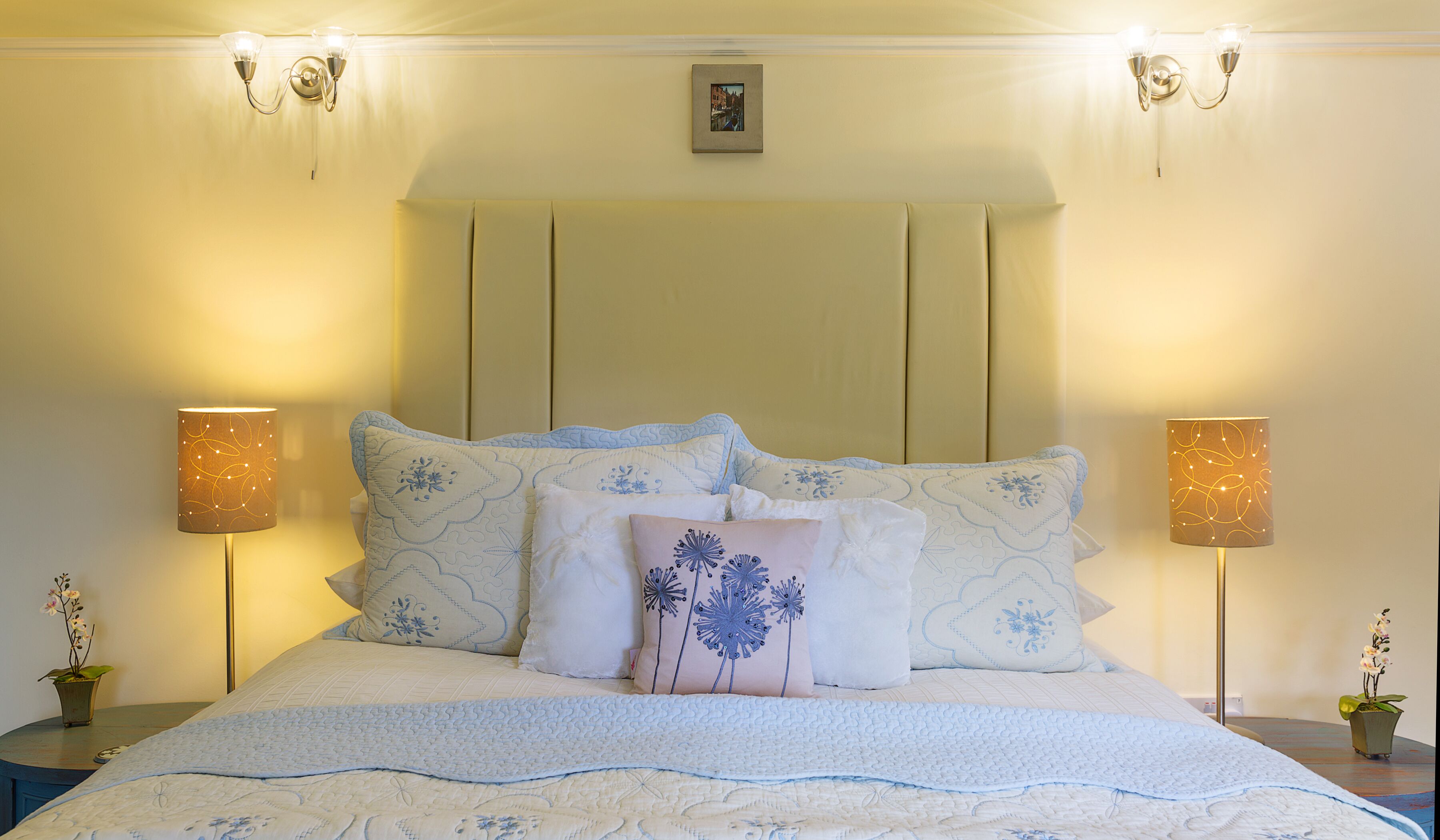 Een netjes opgemaakt bed met een gewatteerde spreidek, met aan weerszijden twee nachtlampjes, en een lijst met een afbeelding boven het hoofdeinde.