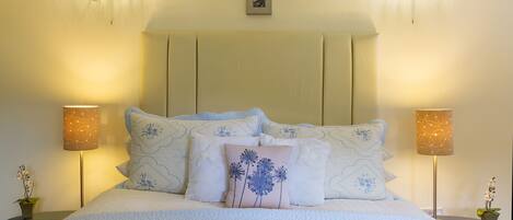 Een netjes opgemaakt bed met een gewatteerde spreidek, met aan weerszijden twee nachtlampjes, en een lijst met een afbeelding boven het hoofdeinde.