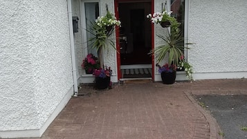 A single-story building with a red door, white exterior walls, and a tiled roof. There are two potted plants on the front porch.