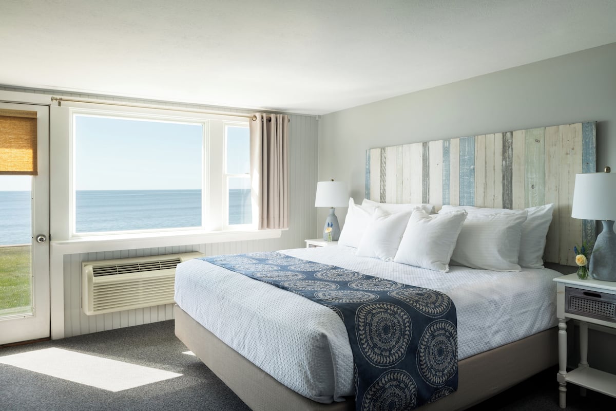 A bedroom with a large window, a bed with a patterned bedspread, two bedside lamps, and a wooden headboard.