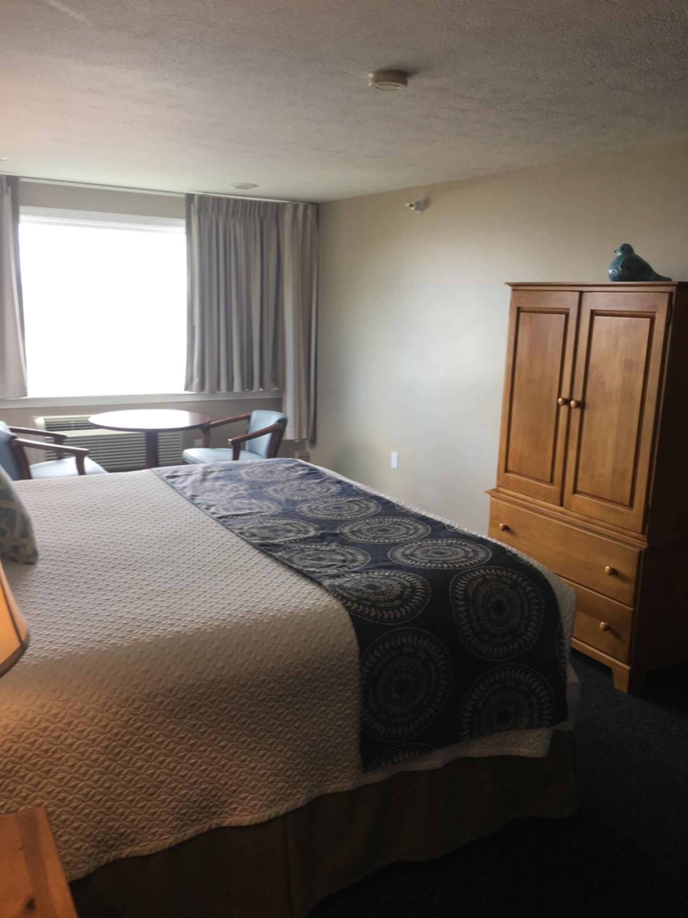 A hotel room with a bed, a wooden wardrobe, a small table, and a window with curtains.