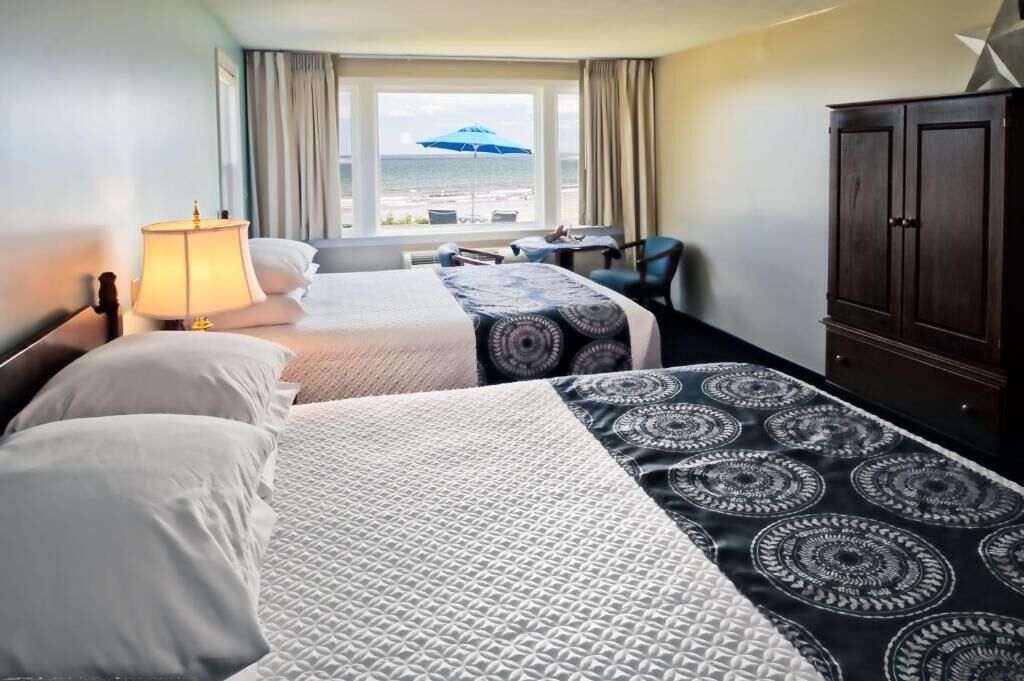 A hotel room with two beds, a desk, a chair, a wardrobe, and a view of the sea through the window.