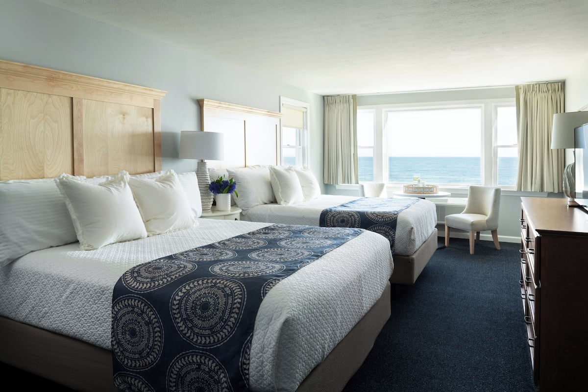 A hotel room with two beds, a desk, a chair, and a view of the sea.
