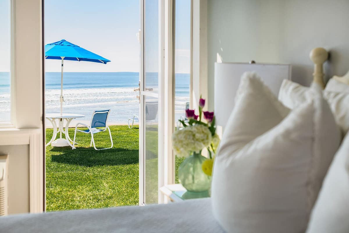 A room with a view of a beach, a blue umbrella, and a white bed with pillows.