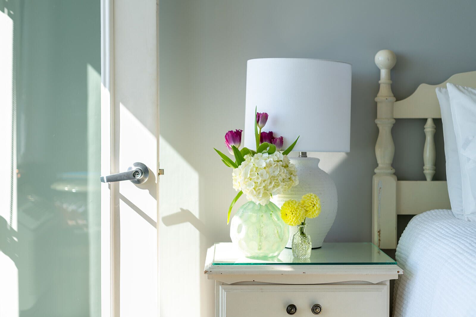 A bedroom with a white bed, a nightstand with a lamp and a vase of flowers, and a door to the left.