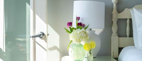 A bedroom with a white bed, a nightstand with a lamp and a vase of flowers, and a door to the left.