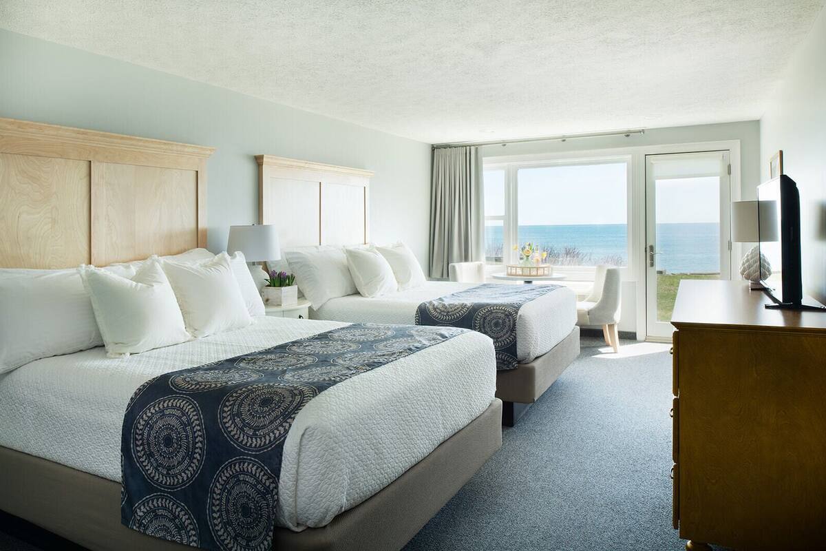 A hotel room with two beds, a desk, a chair, a TV, and a view of the sea.