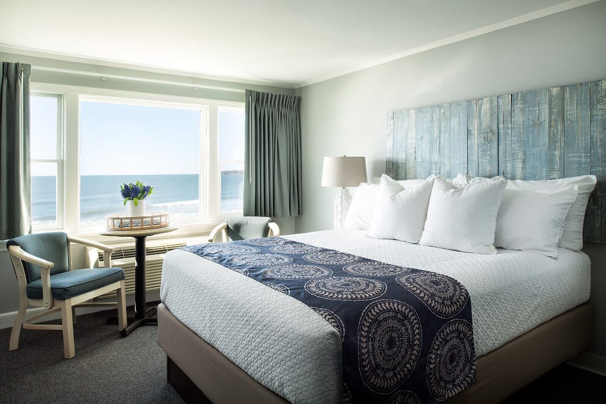 A hotel room with a large bed, a desk with a chair, a view of the ocean, and a lamp.