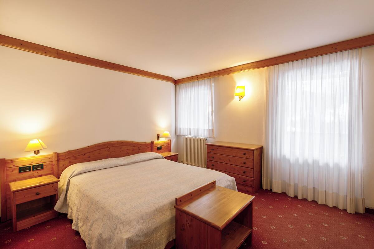 Ein Hotelzimmer mit einem hölzernen Bett, Nachttischen mit Lampen, einer Holzkommode und einem Fenster mit transparenten Vorhängen.