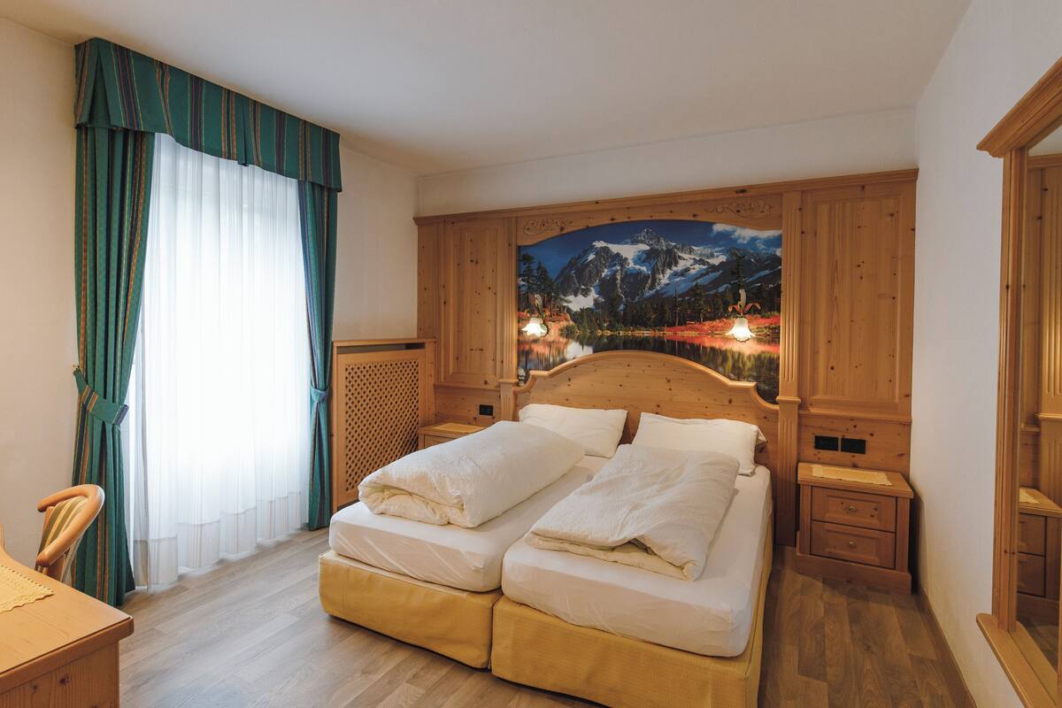 Ein Schlafzimmer mit einem Doppelbett, einem Holz-Kopfteil, einem Nachttisch, einem Schreibtisch und einem großen Fenster mit Vorhängen.