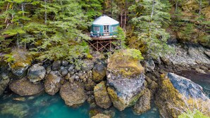 A cabin on stilts surrounded by dense forest and rocky shoreline. - Orca Island Cabins (Seward)