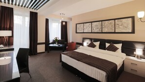 Habitación de hotel con una cama grande, un escritorio, una silla, una mesita con un florero y una pared decorada con obras de arte enmarcadas.