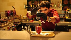 Un cantinero preparando una bebida en un bar, con estantes llenos de botellas y una pared decorativa.