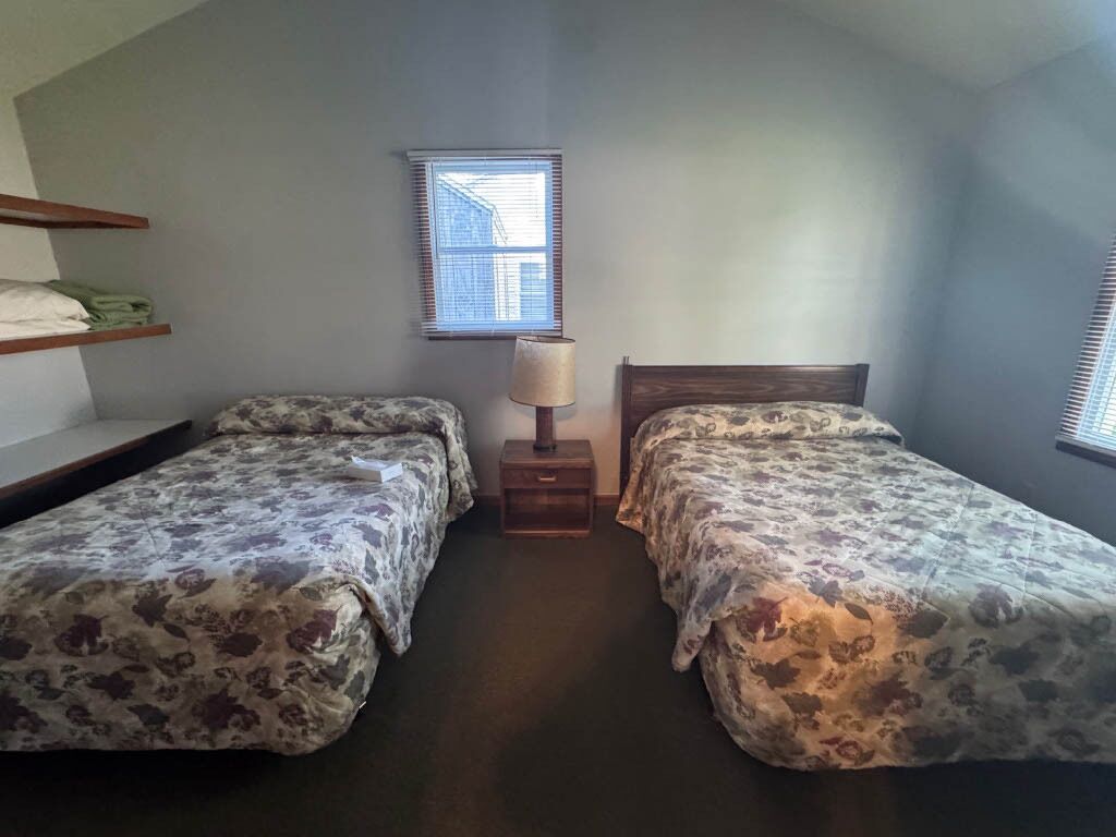 A bedroom with two beds, a nightstand, a lamp, and a window.