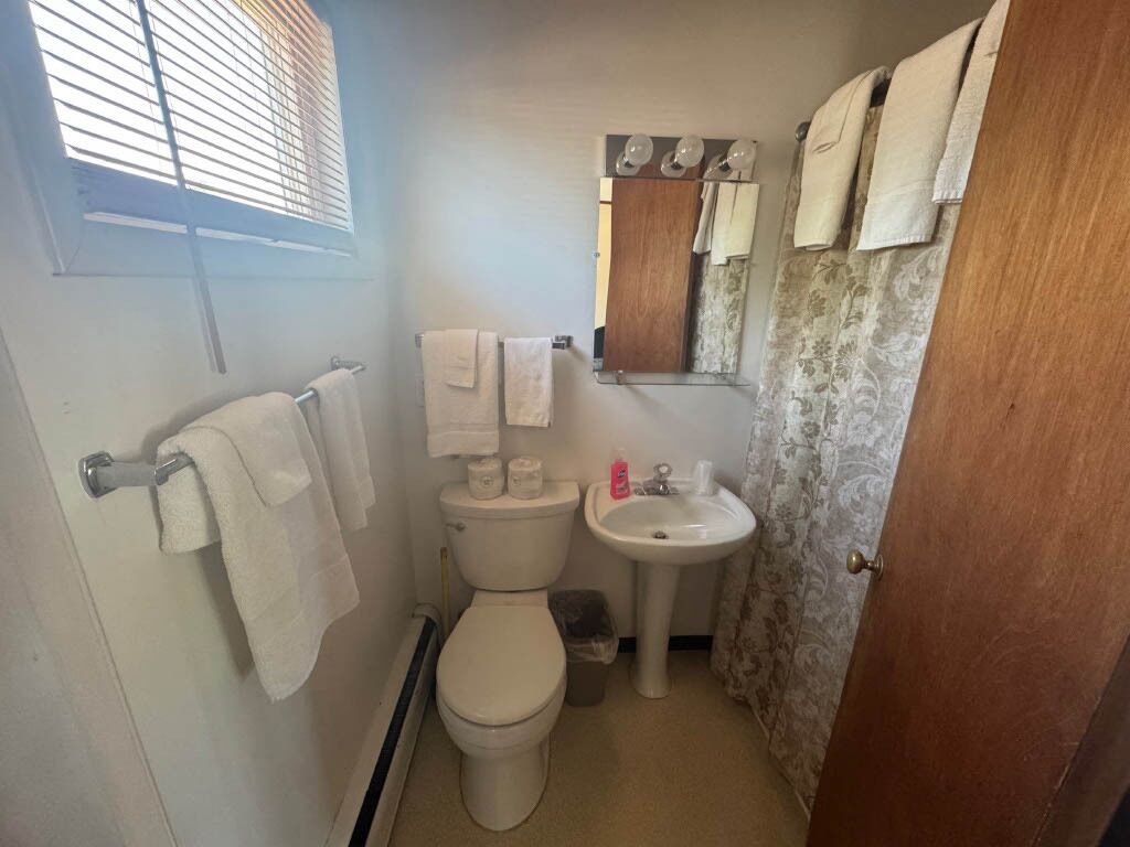 Un baño pequeño con inodoro, lavamanos y un espejo sobre el lavamanos. Hay toallas colgadas en la pared y una cortina de ducha.