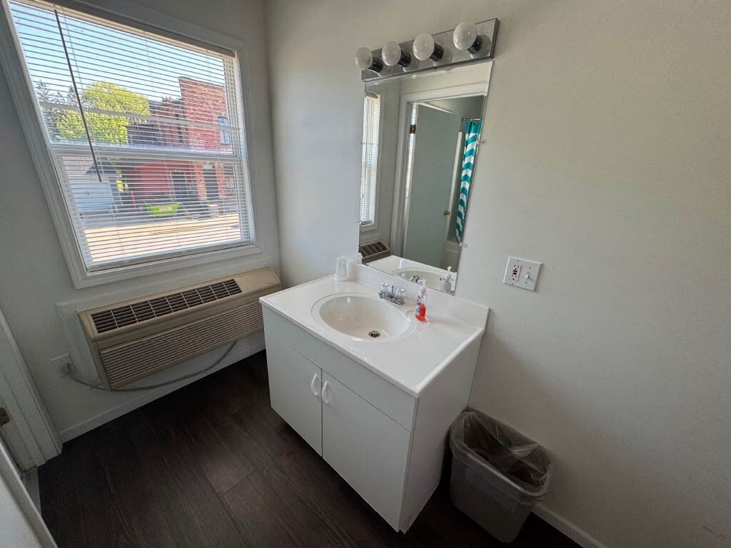 A bathroom with a white sink, mirror, and vanity. There is a window with blinds, a vent, and a trash can.